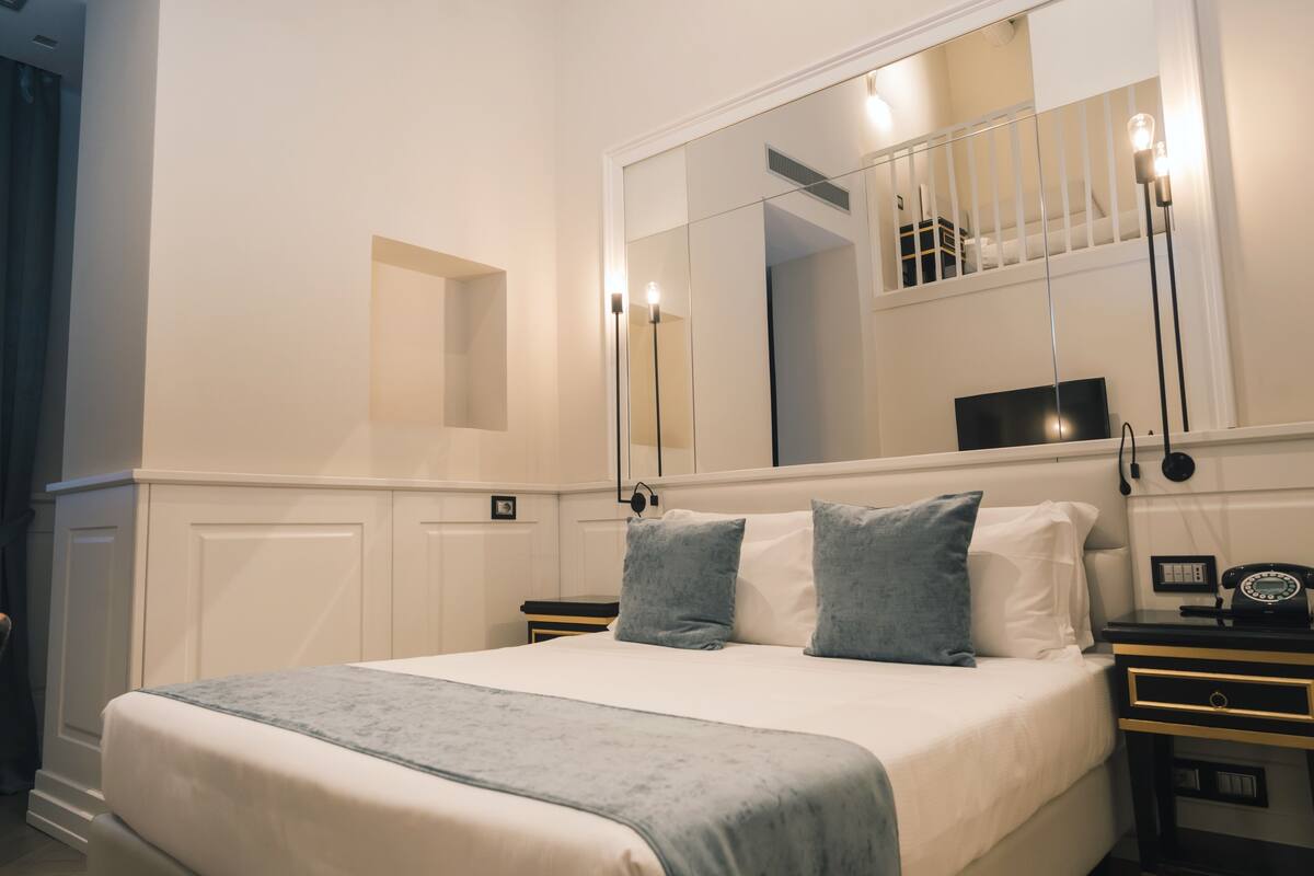 Un letto rifatto con cuscini, un comodino con una lampada e uno specchio grande sopra il letto.