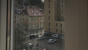 Blick durch ein Fenster auf einen Parkplatz mit mehreren Autos, einem Fußgänger und Gebäuden im Hintergrund.