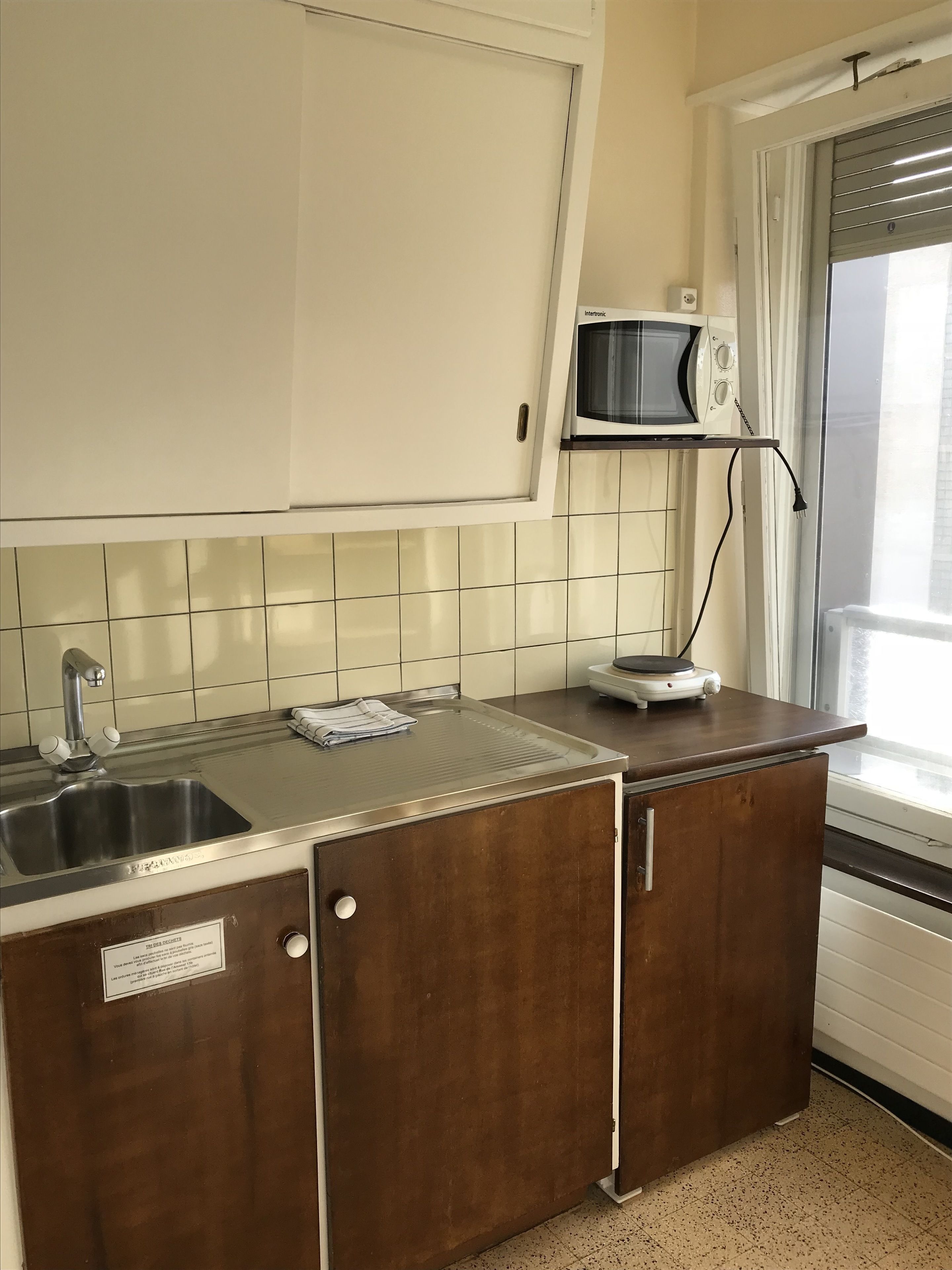 A kitchen with a stainless steel sink, a microwave on a shelf, and a window with curtains.