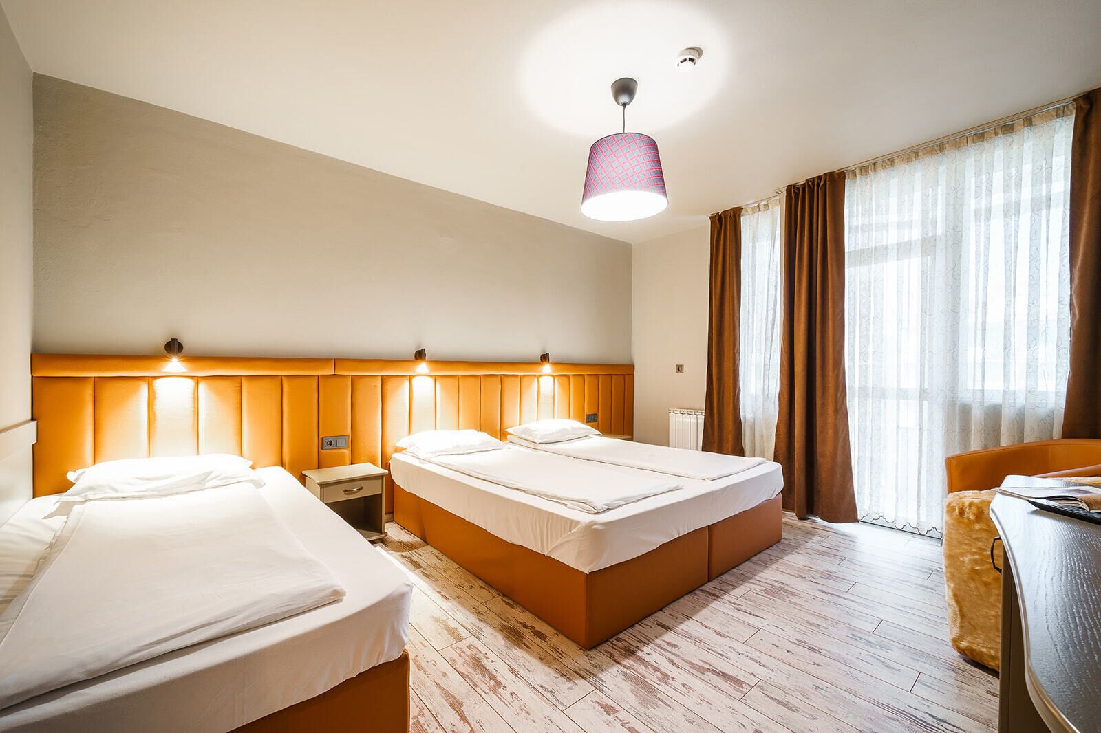 A hotel room with two beds, a wooden headboard, a desk, and a large window with curtains.