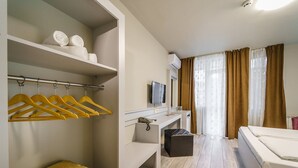 Habitación de hotel con cama, escritorio, televisor, espejo, armario con perchas y cesto de basura.