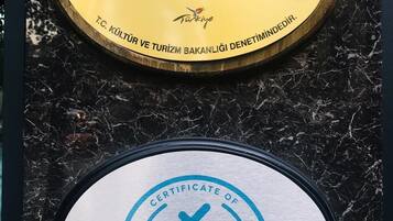 Two plaques on a marble surface. The top plaque is gold with the text "YENİ BAHAR OTEL" and a Turkish flag logo. The bottom plaque is white with "CERTIFICATE OF SAFE TOURISM" and "Yeni Bahar Otel" text, along with logos for TUV Austria and Turkey.