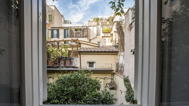 A view through a window of a narrow urban street with buildings, a small tree, and a metal grate at the window base. - Trevi Contemporary Suite (Rome)