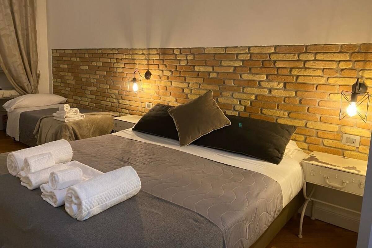 A hotel room with a brick headboard, a bed with grey bedding, two bedside tables, and a suitcase on one table.