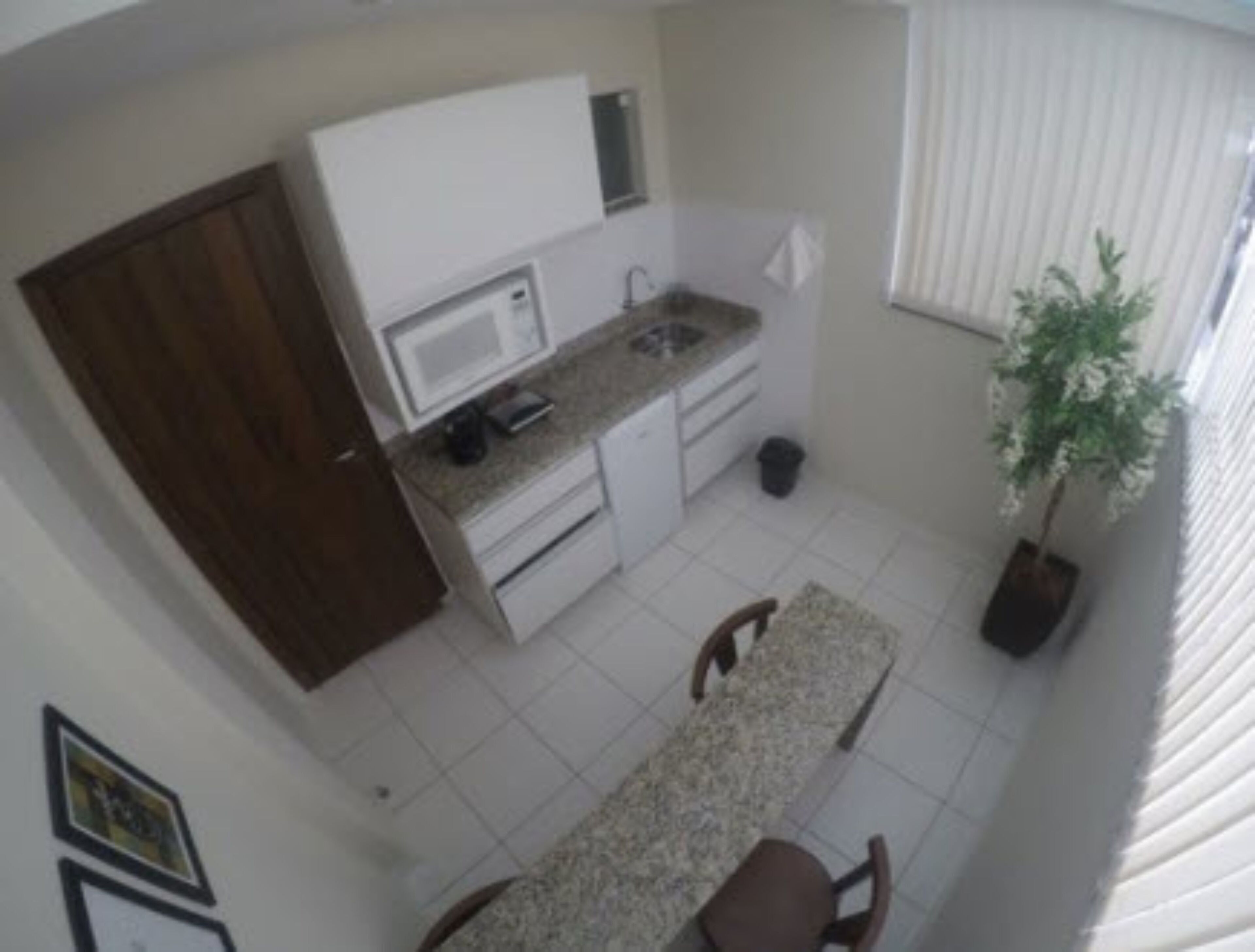 A compact kitchen with white cabinets, a microwave, and a sink. There's a dining table with chairs, a potted plant, and a window with blinds.