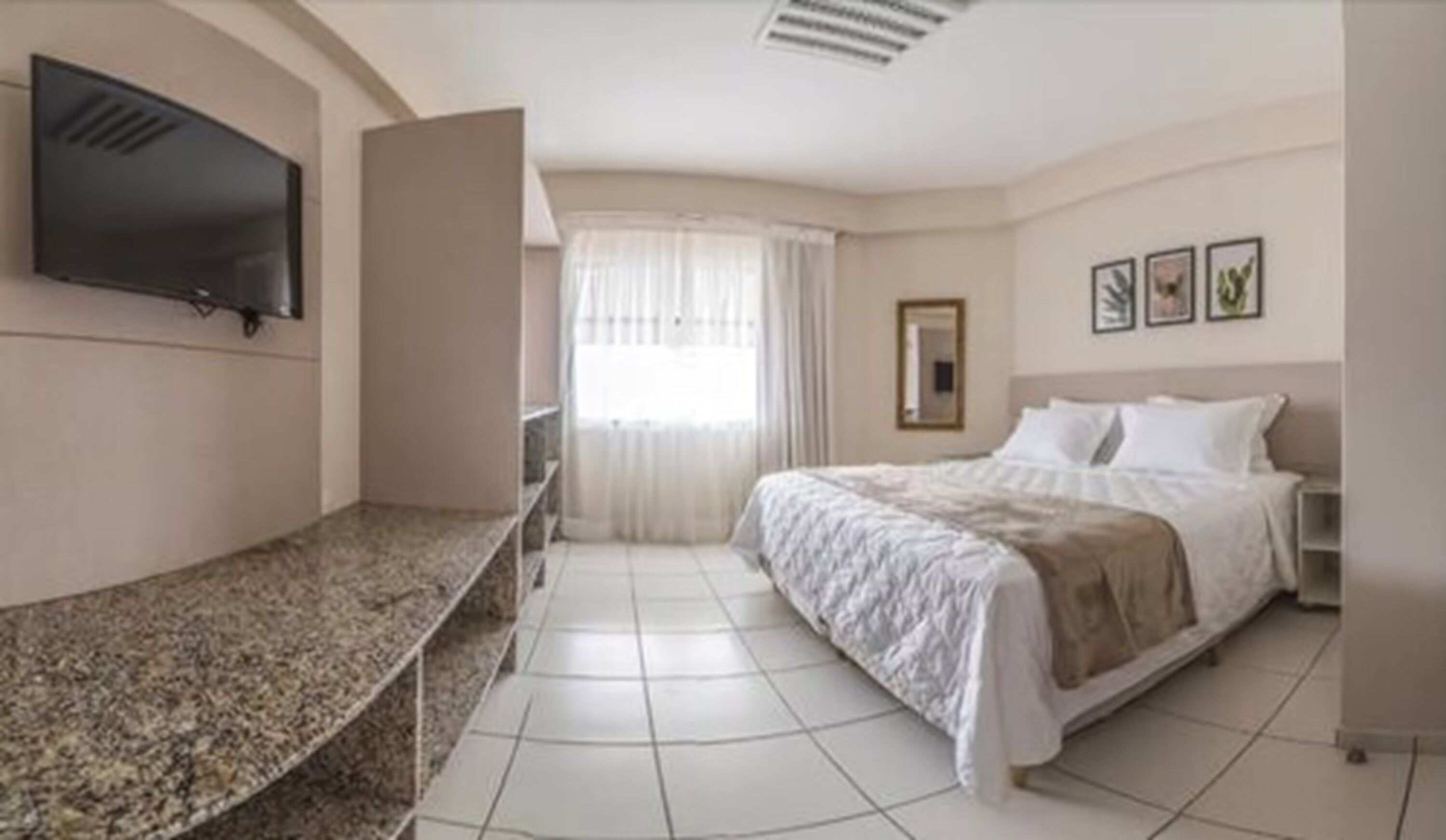 A hotel room with a bed, a TV mounted on the wall, a window with curtains, and a tiled floor.
