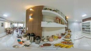 A spacious dining area with multiple tables, a central serving station, and a kitchen area with shelves above. - Pousada Doluar (Penha)