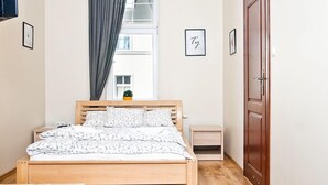A bedroom with a wooden bed, a bedside table, a small bedside table, a dark rug, a wooden bench, a window with curtains, a door, and a wall-mounted light fixture. - Smart Rooms for Rent (Kraków)