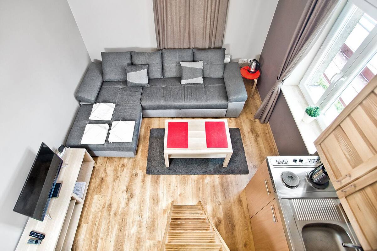 Un espacio habitable compacto con un sofá gris, una mesita de madera, una cocina con estufa y fregadero, y un ventanal con cortinas.