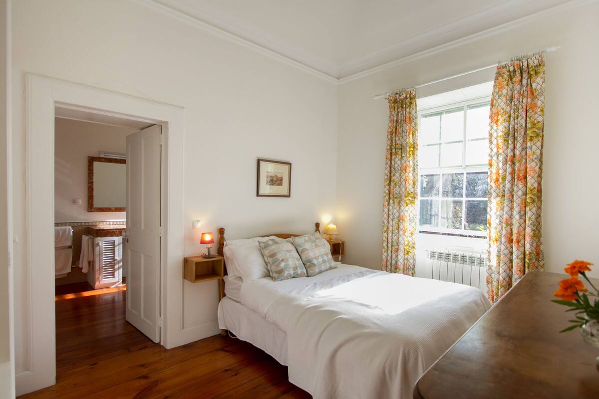 A bedroom with a bed, bedside lamps, a window with curtains, a picture on the wall, and a wooden floor.