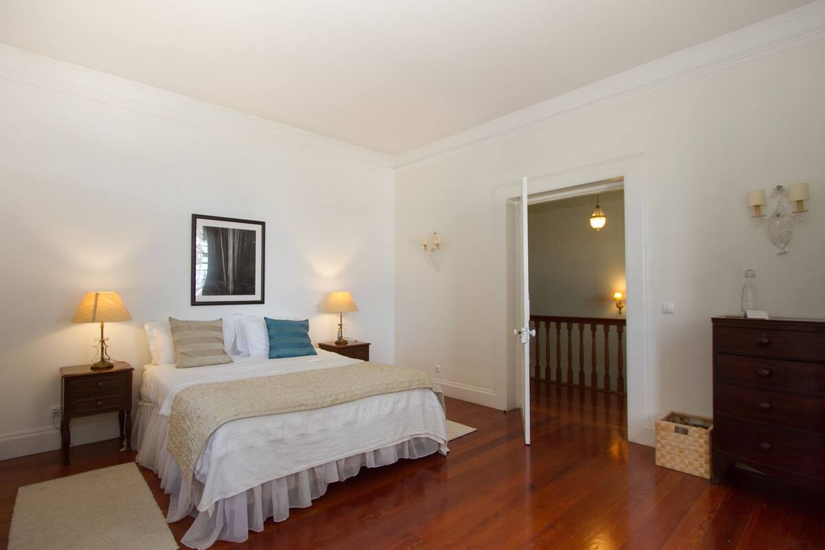 A bedroom with a bed, two bedside lamps, a framed picture, a dresser, and a door leading to another room.