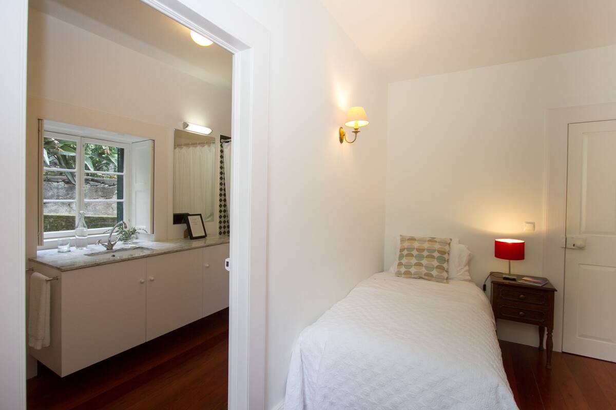 A bedroom with a bed, a nightstand, a bedside lamp, a window with curtains, a mirror, and a view of the outside.