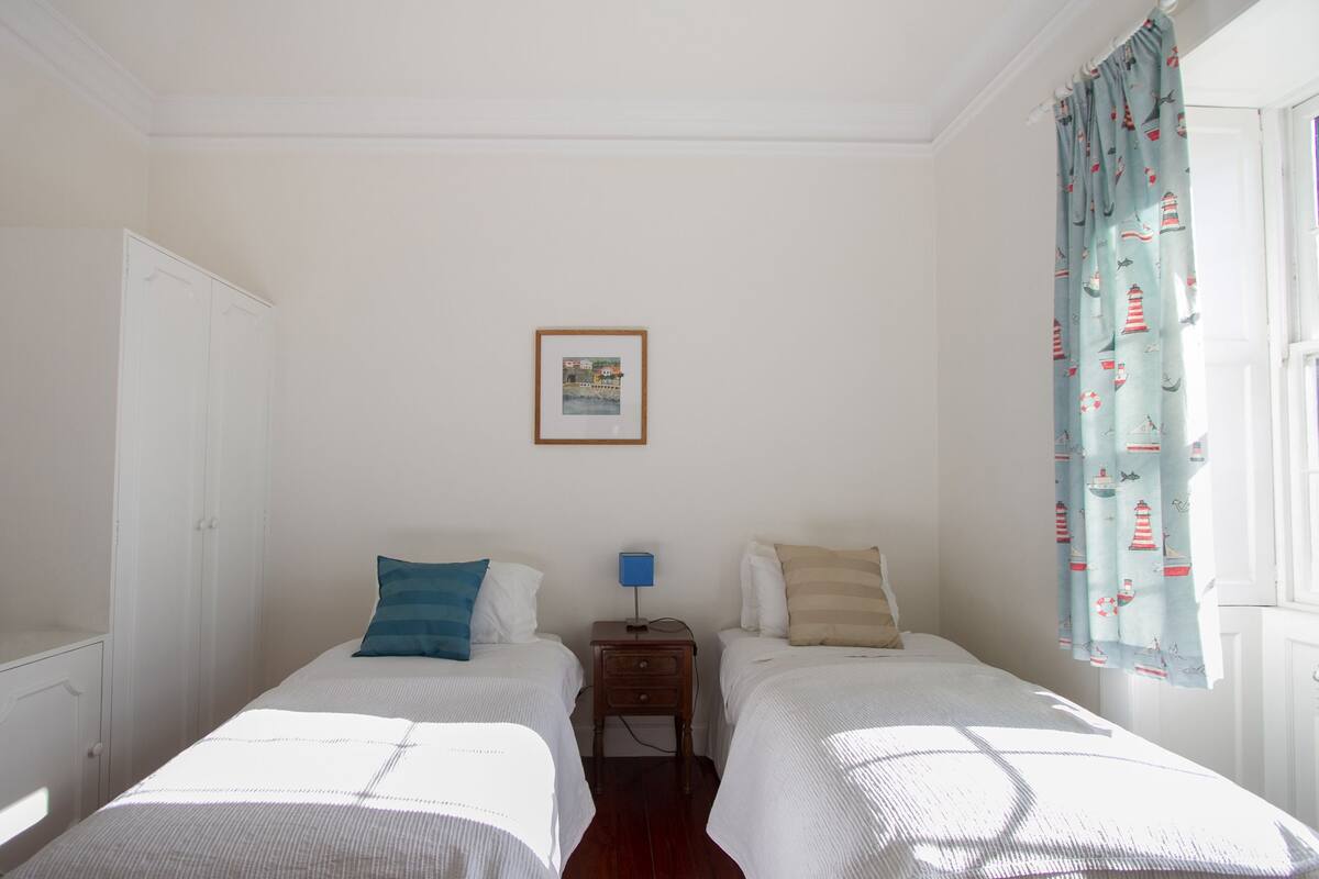 A room with two beds, a nightstand with a lamp, a framed picture on the wall, and a window with curtains.
