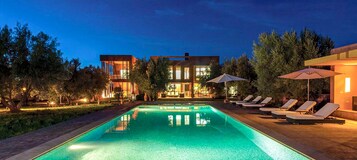Uma área de piscina moderna com espreguiçadeiras e guarda-sóis, rodeada por árvores e um edifício com grandes janelas.