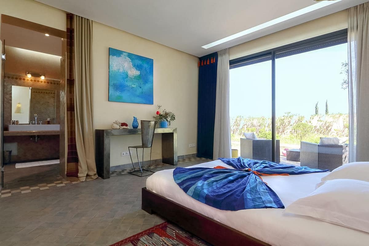 A hotel room with a large bed, a desk, a chair, a bathroom, and a balcony with a view.