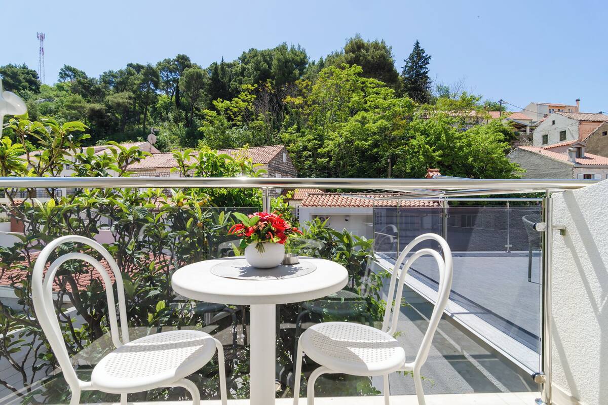 Un balcon avec une table ronde et deux chaises, un pot de fleurs aux fleurs rouges, et une vue sur une ville avec des bâtiments aux toits de tuiles.