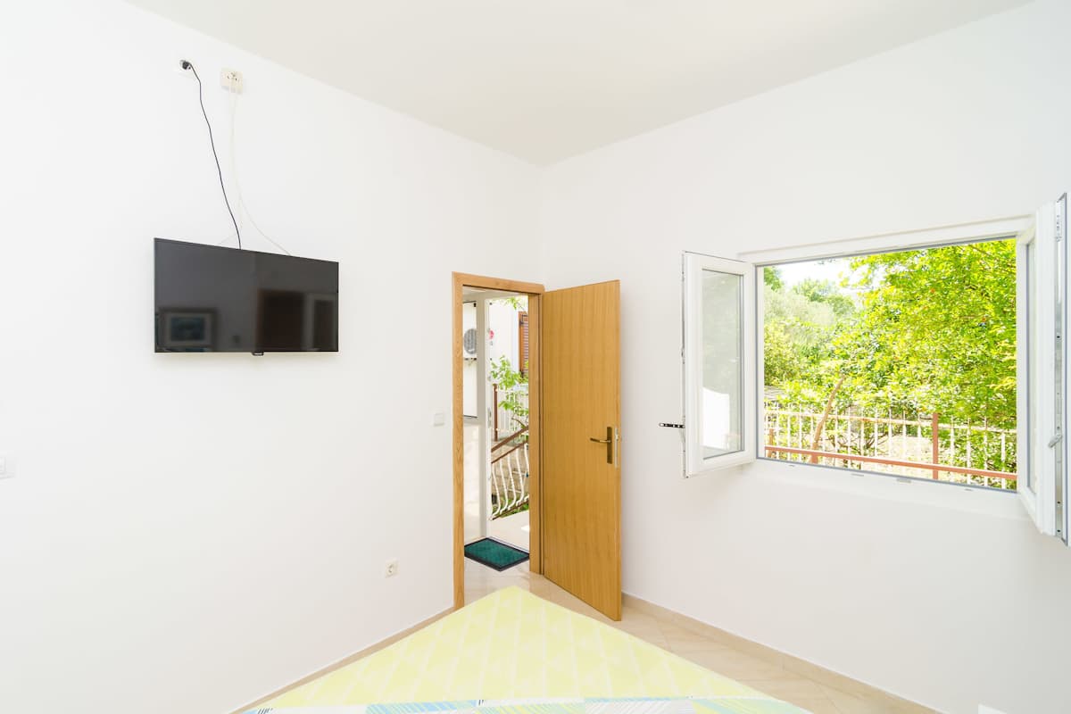 Une chambre dotée d’un téléviseur à écran plat fixé au mur, d’une porte en bois donnant sur un balcon et d’une grande fenêtre offrant une vue sur la verdure.