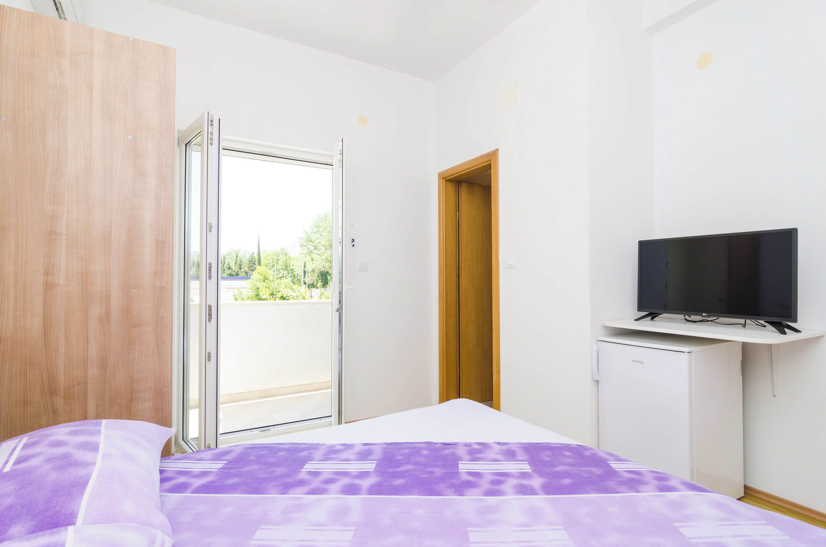 A bedroom with a bed, a television on a stand, a wardrobe, and a sliding glass door leading to a balcony.