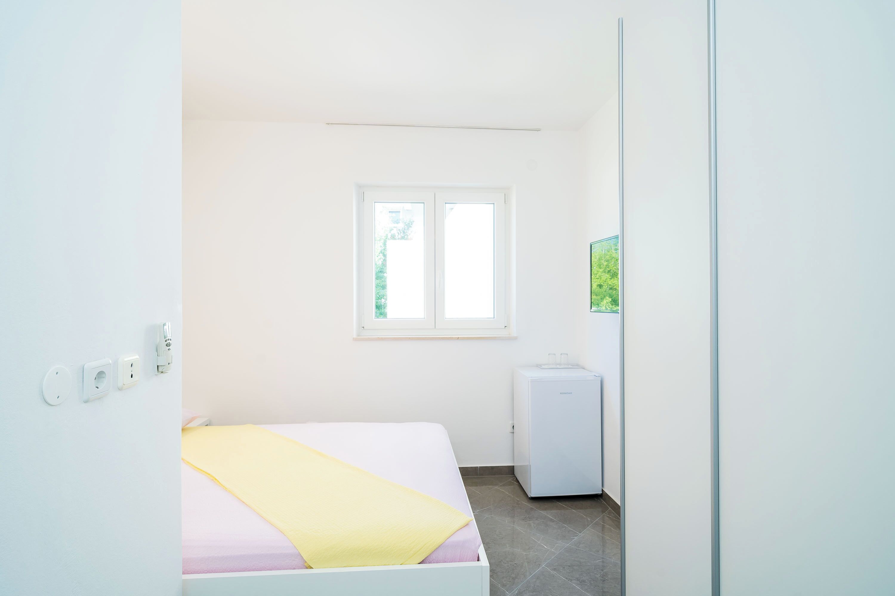 Una habitación pequeña y luminosa con una cama, un ventanal y un electrodoméstico blanco compacto.