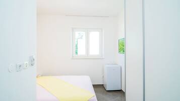 Una habitación pequeña y luminosa con una cama, un ventanal y un electrodoméstico blanco compacto.