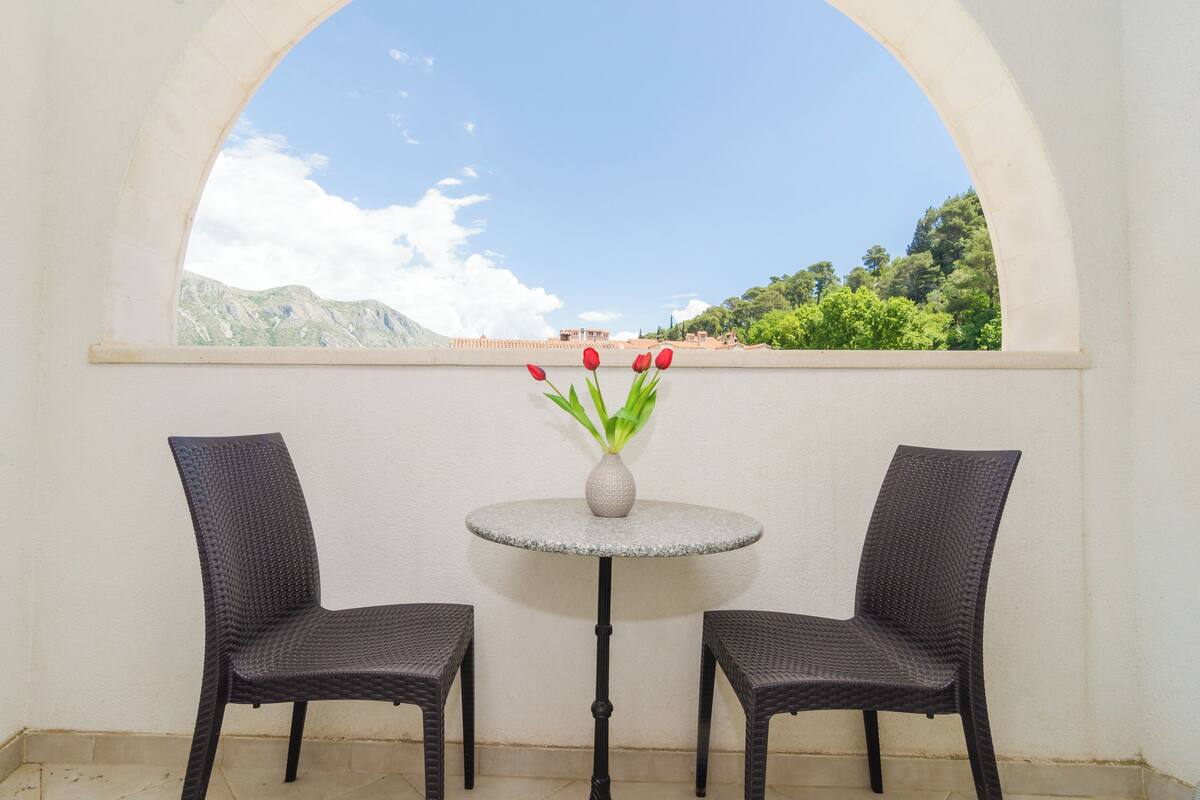 Un balcon avec deux fauteuils en osier et une petite table ronde sur laquelle est posé un vase rempli de fleurs rouges.