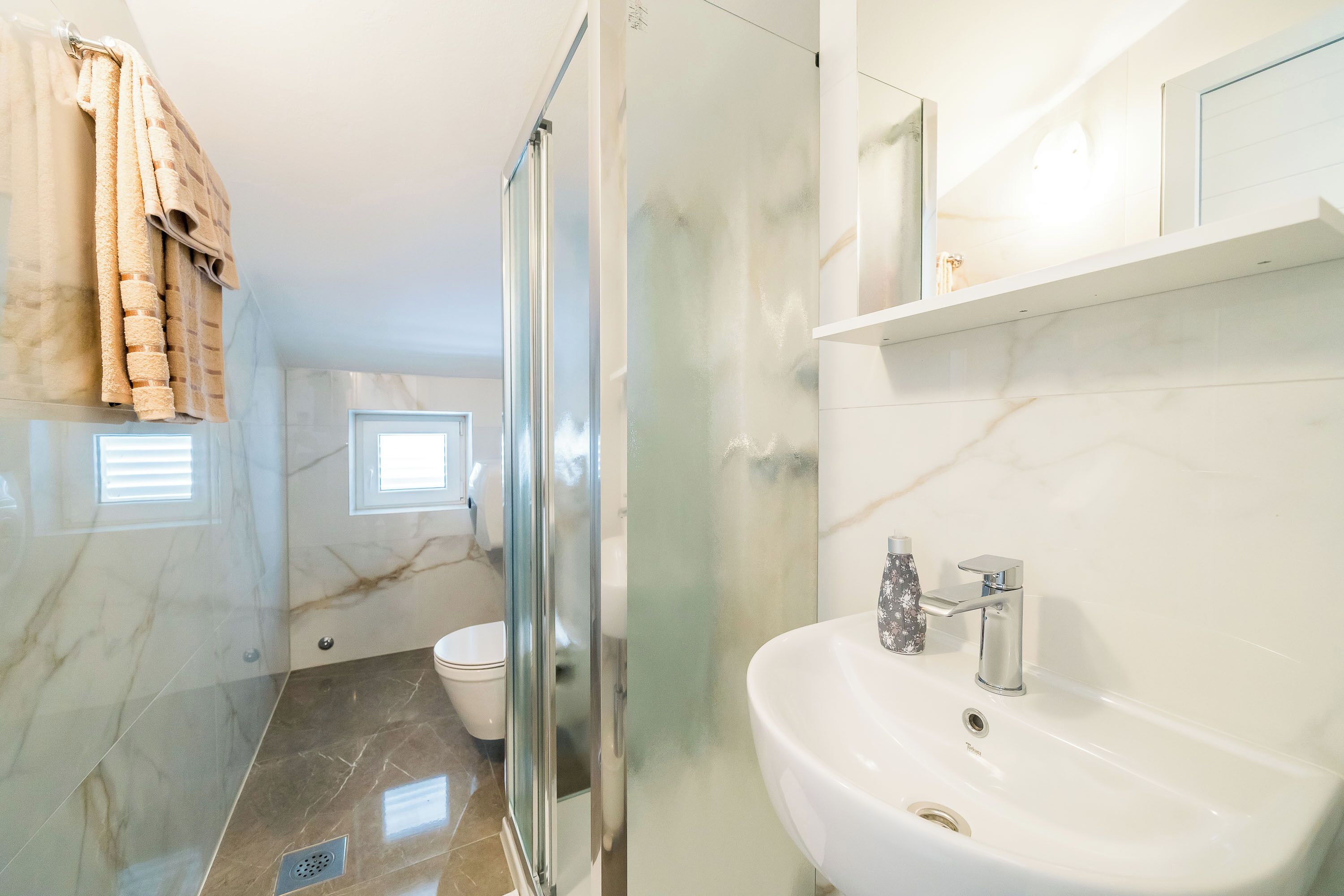 A bathroom with a glass shower enclosure, a white sink, and a marble-tiled floor.