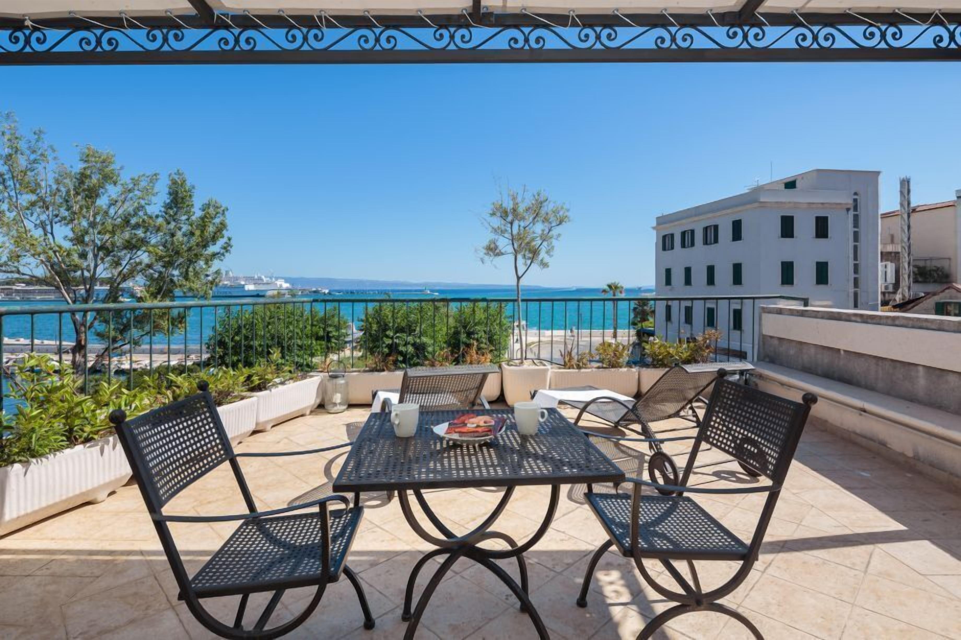 A terrace with a table and chairs, overlooking a body of water and buildings.