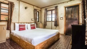 Habitació d'hotel amb un llit gran, panell de fusta al capçal, dues finestres amb cortines, una porta de fusta i un paviment de rajoles.