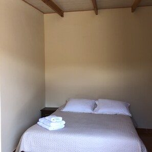 A single bed with a white quilt and pillows, a small bedside table, and a stack of folded white towels.