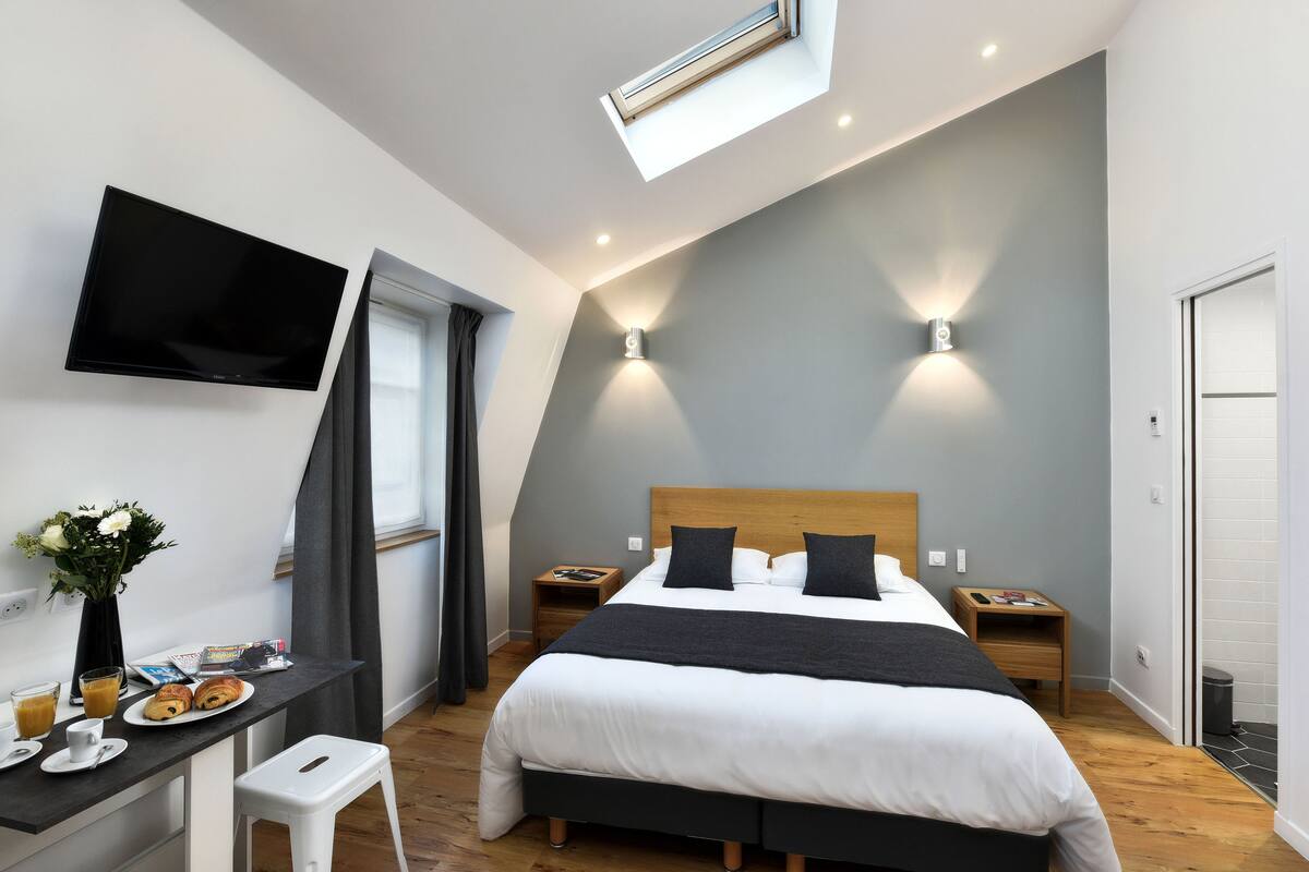 Un dormitorio moderno con cama, mesitas de noche, un escritorio con silla, televisor de pantalla plana y tragaluz.