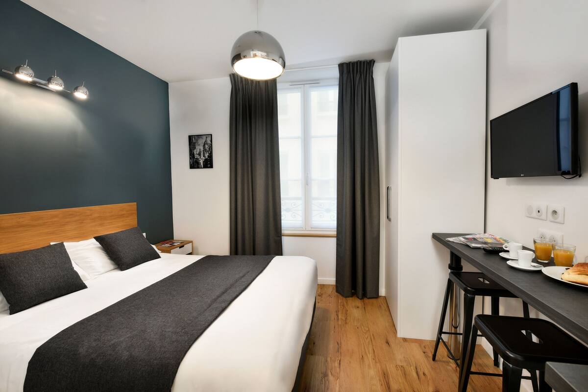 Una habitación de hotel moderna con una cama, un escritorio con silla, un televisor y un ventanal con cortinas.