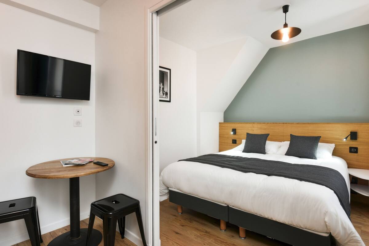 Un dormitorio moderno con una cama, un televisor montado en la pared, una mesita redonda y techo de madera.