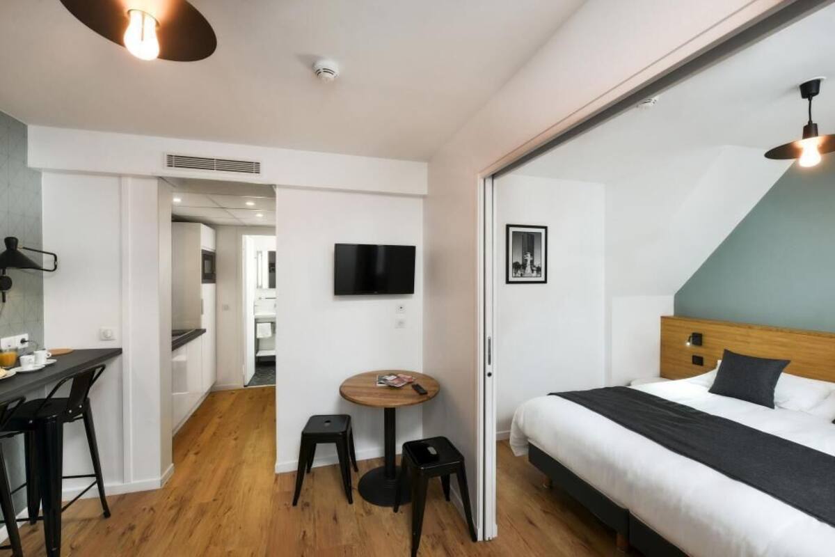 Una habitación de hotel moderna con una cama, un televisor, una zona de comedor con mesa y sillas, y un taburete pequeño.
