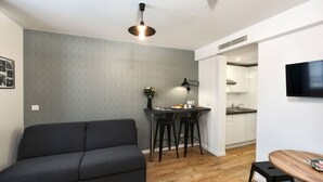 A modern living space with a grey sofa, a dining area with two black chairs, a wooden table, and a kitchen in the background. - Hôtel Résidence Montebello (Paris)