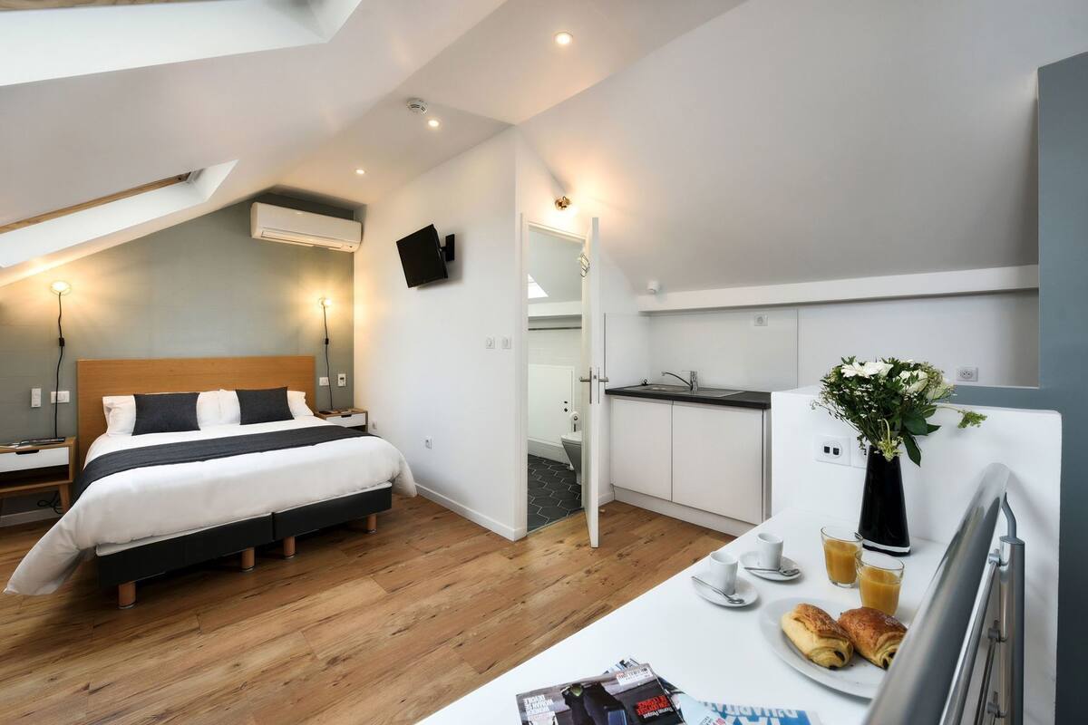 Una habitación de hotel moderna con cama, mesita de noche, pequeño refrigerador, lavabo y una pequeña mesa de desayuno.