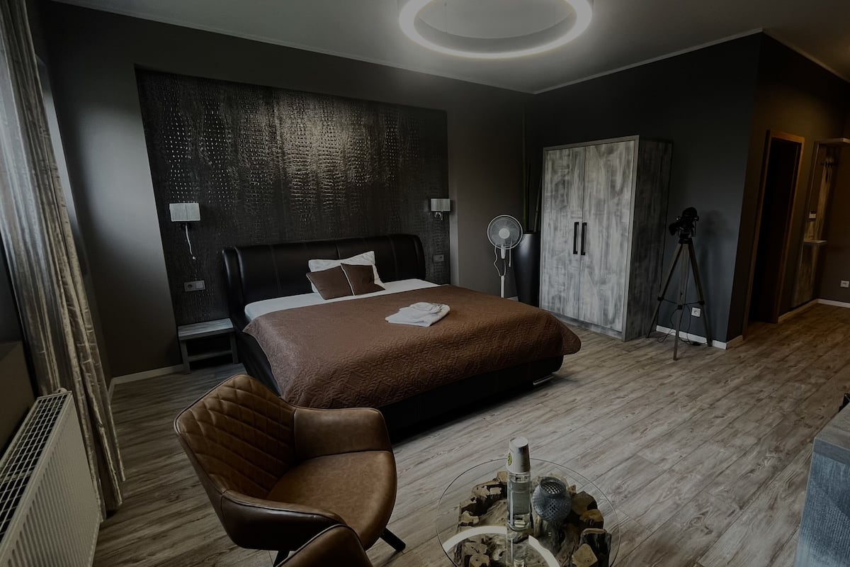 A modern hotel room with a large bed, a sitting area with a chair, a desk, and a wardrobe.