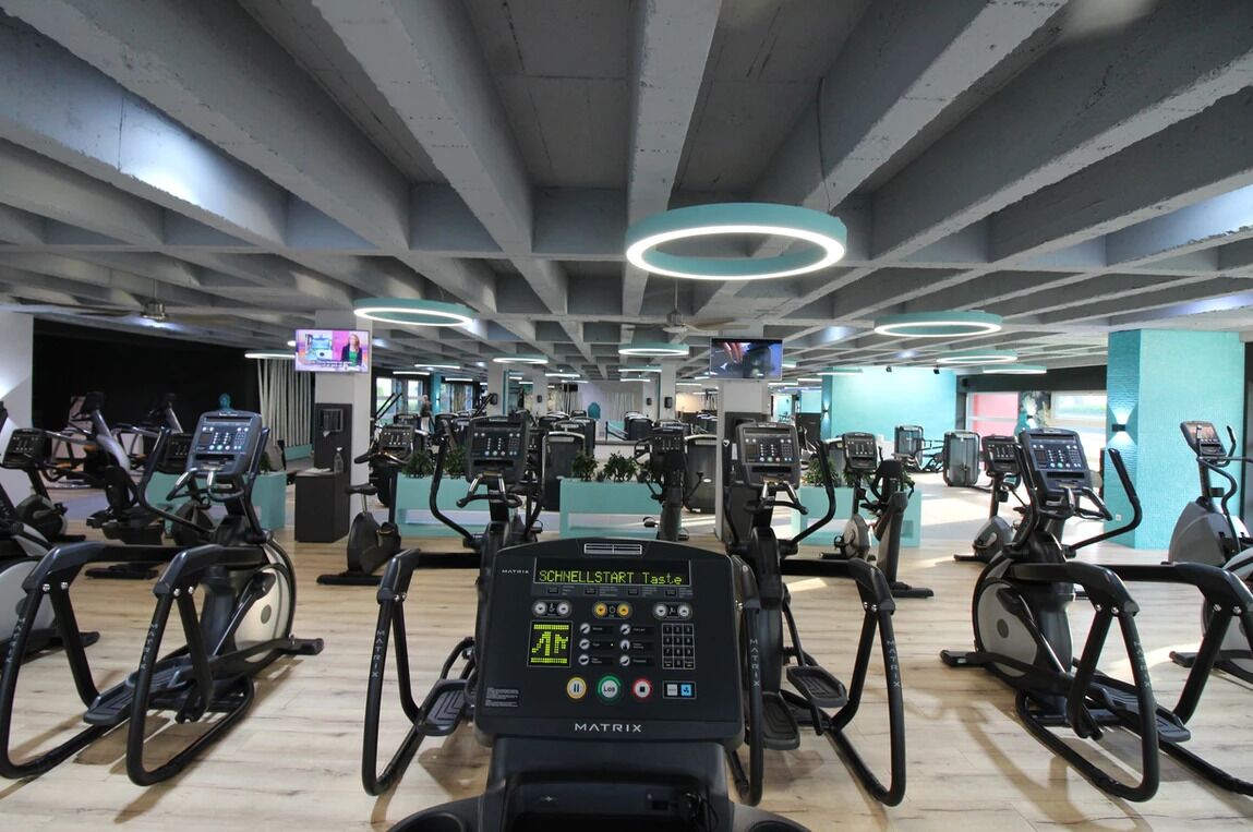 Ein Fitnessstudio mit mehreren Ergometer und Laufbändern, einem modernen Deckengestaltungsstil und einem sichtbaren Fernsehbildschirm.
