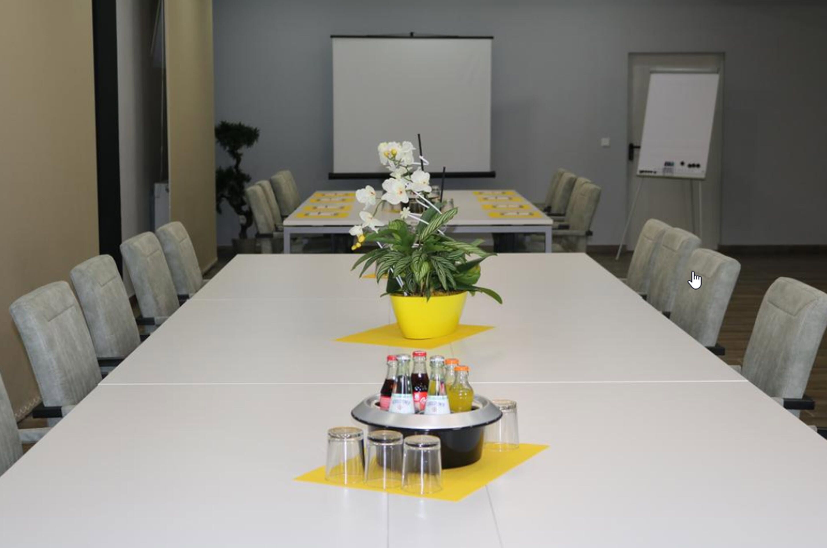 A conference room with a long table, chairs, a projector screen, a whiteboard, and a small plant.
