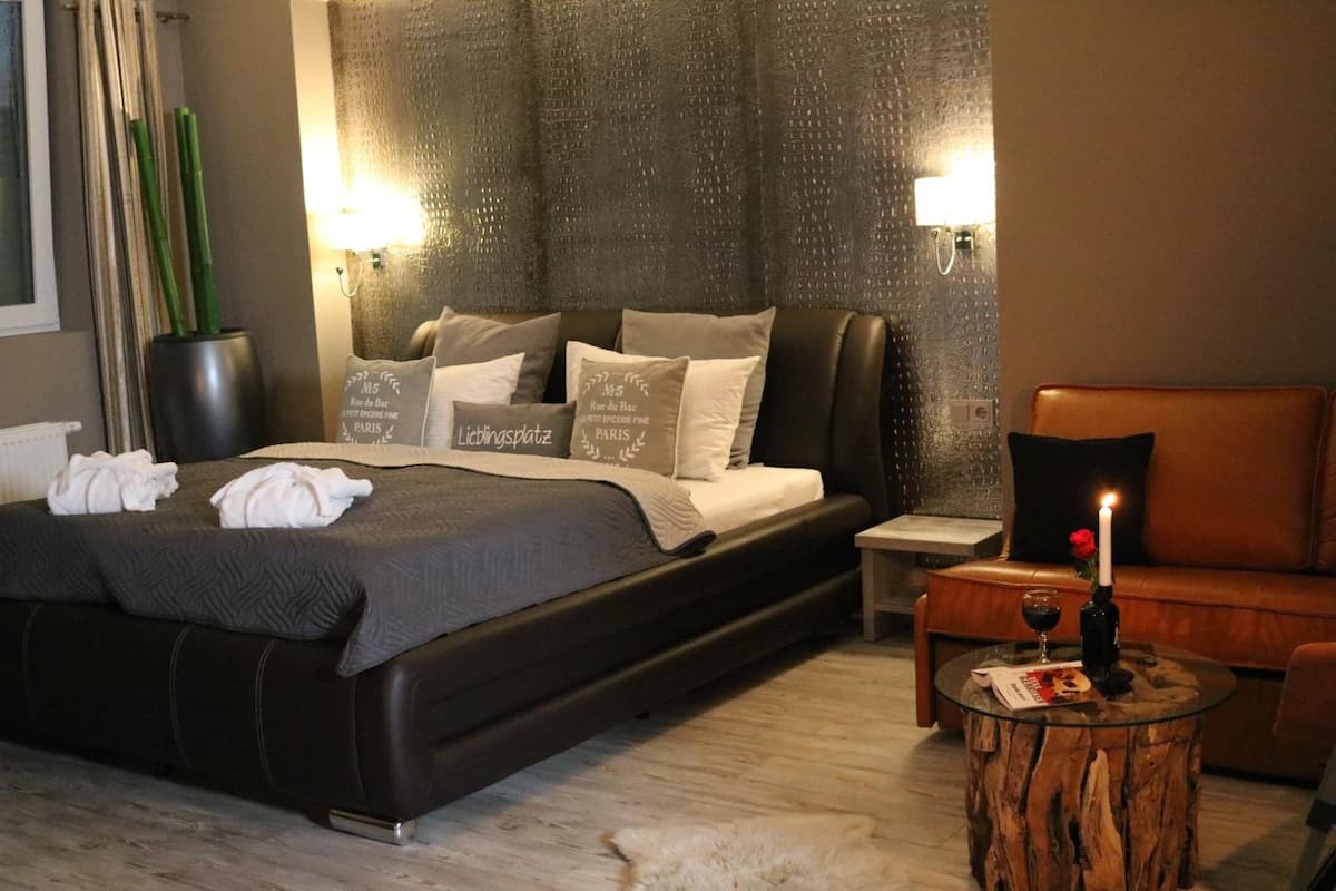 A modern hotel room with a large bed, a wooden side table, a glass coffee table, and a leather armchair.