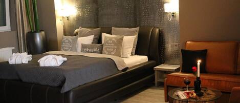 A modern hotel room with a large bed, a wooden side table, a glass coffee table, and a leather armchair.