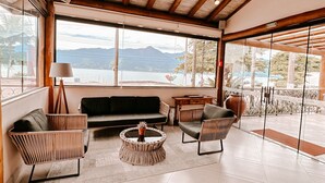 A spacious room with a high ceiling, large windows, and a view of mountains. There are several seating areas, including a sofa, chairs, and a round table. - Abricó Beach Hotel (São Sebastião)