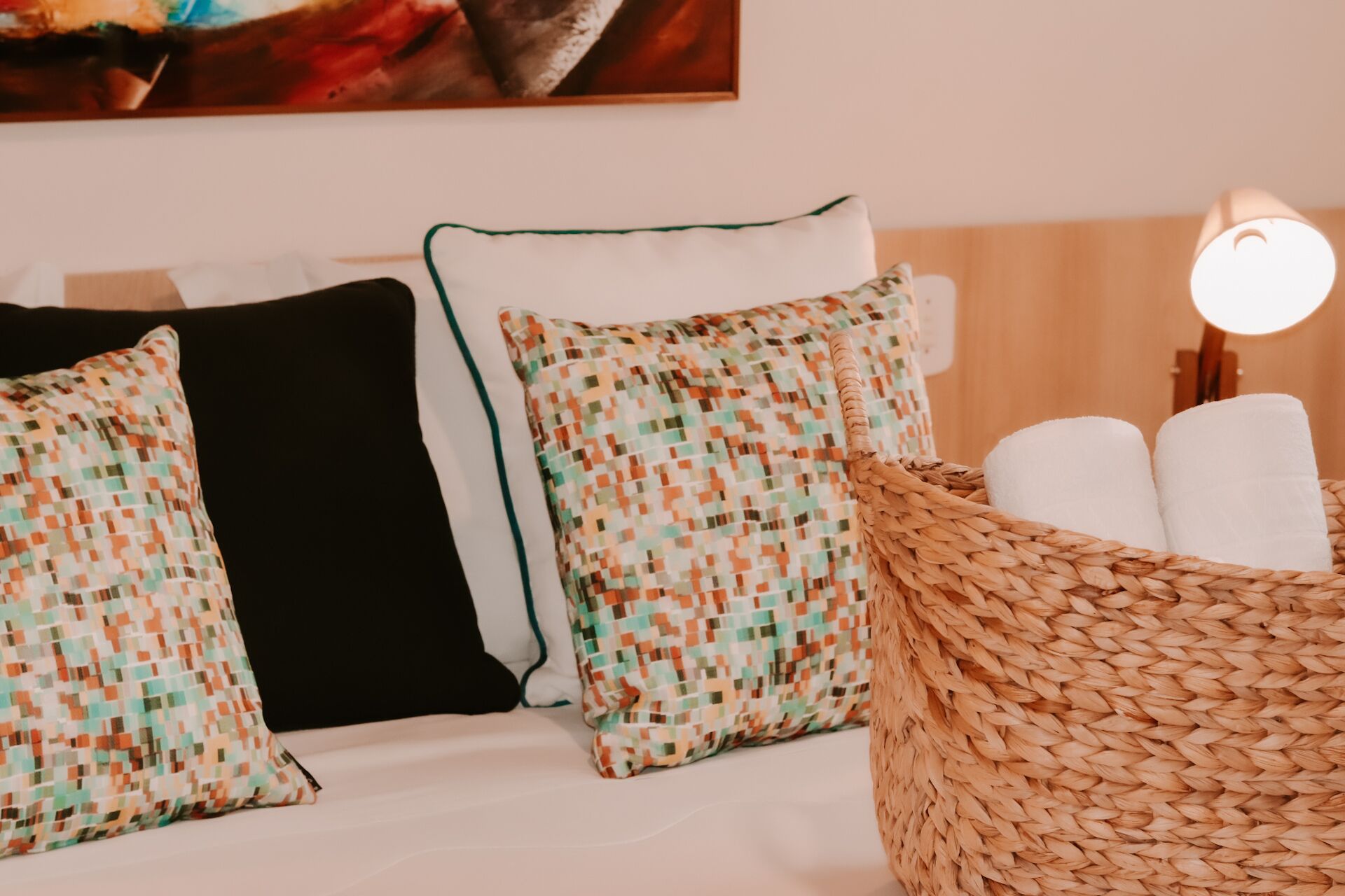 A hotel room with a bed, pillows, a woven basket with towels, and a wall-mounted lamp.