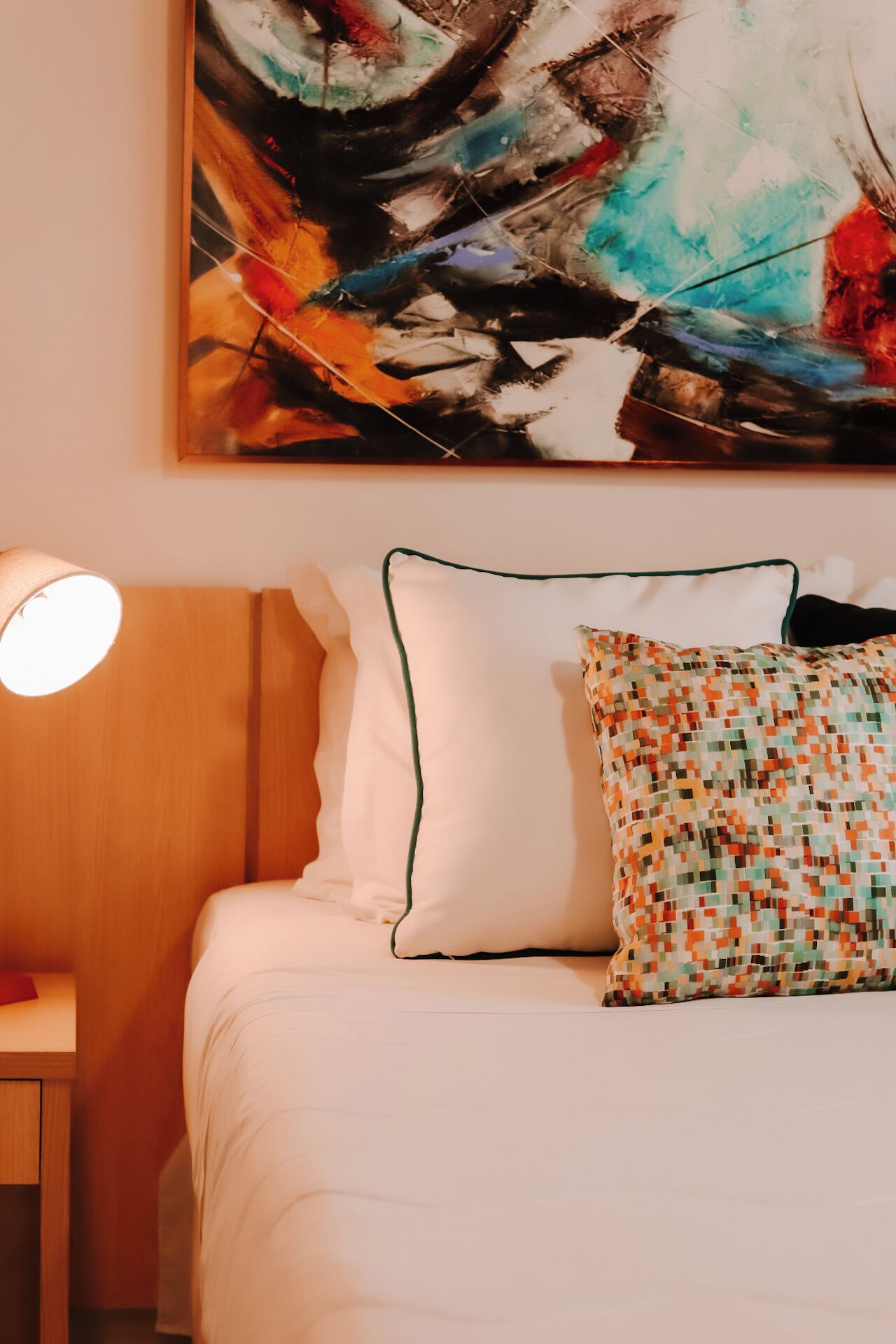 A neatly made bed with white bedding and two pillows, one plain and one with a colorful pattern. A round table lamp is on the left side of the bed.