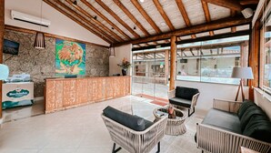 A reception area with a wooden ceiling, a stone wall, a colorful lion painting, wicker furniture, and a view through large windows. - Abricó Beach Hotel (São Sebastião)