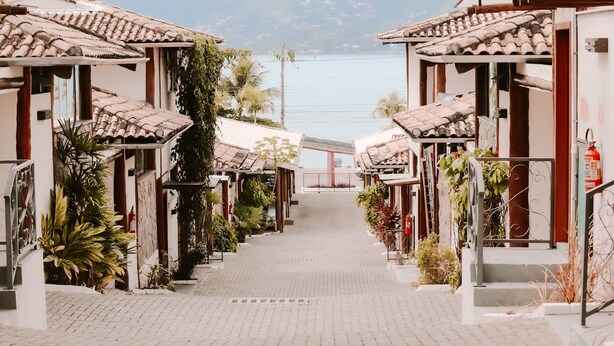 A row of traditional-style buildings with tiled roofs, a paved pathway, and a view of a body of water in the distance. - Abricó Beach Hotel (São Sebastião)