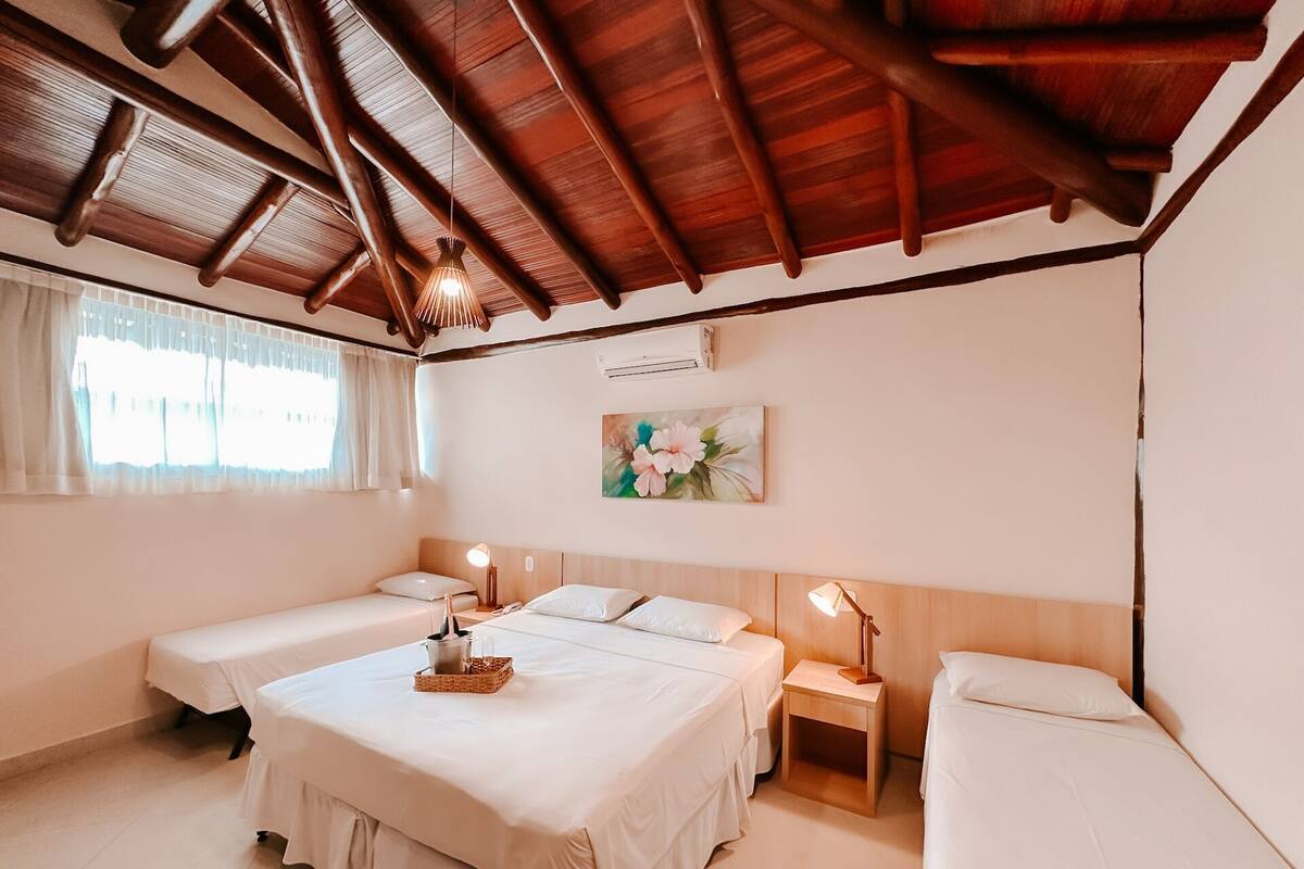 A hotel room with a wooden ceiling, two single beds, a large bed, a small bedside table with a lamp, a painting on the wall, and a window with curtains.