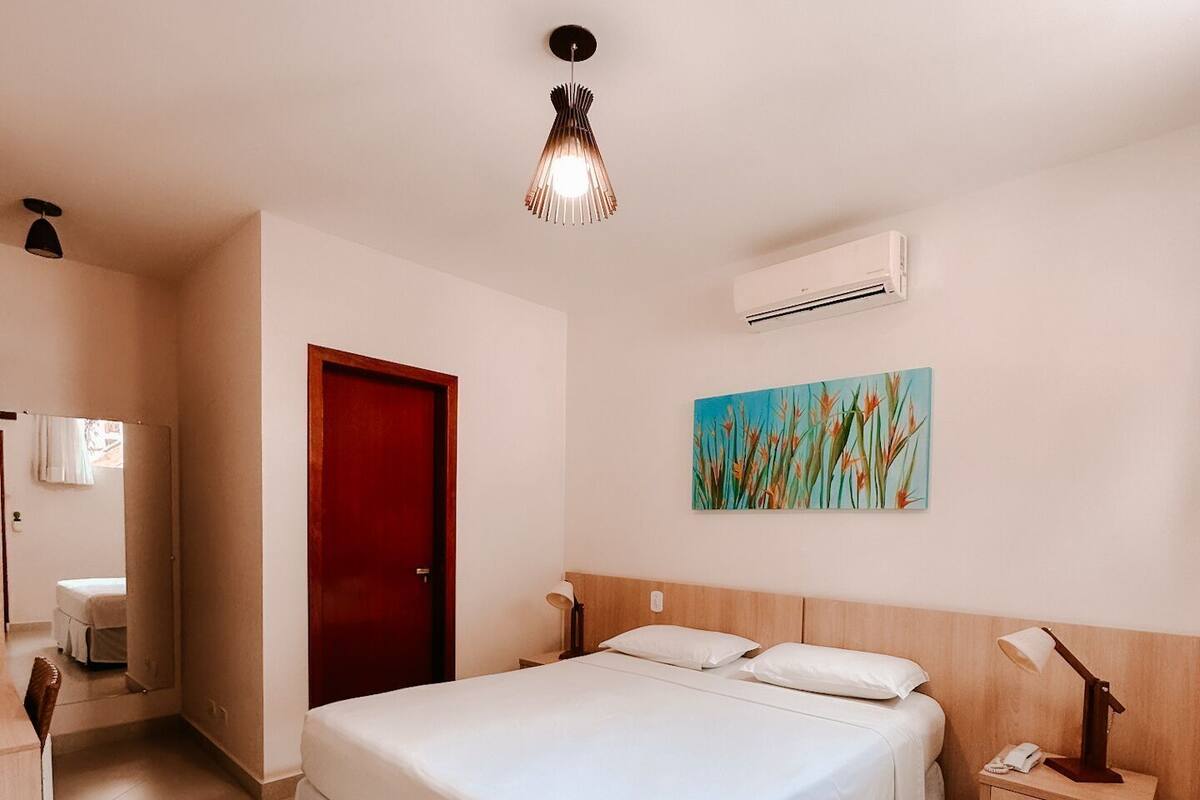 A hotel room with a large bed, a wooden headboard, a nightstand with a lamp, an air conditioning unit, and a painting on the wall.