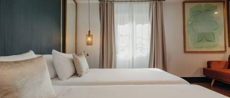 Habitación de hotel con cama, una silla, ventana con cortinas, lámpara de pared y una obra de arte enmarcada.