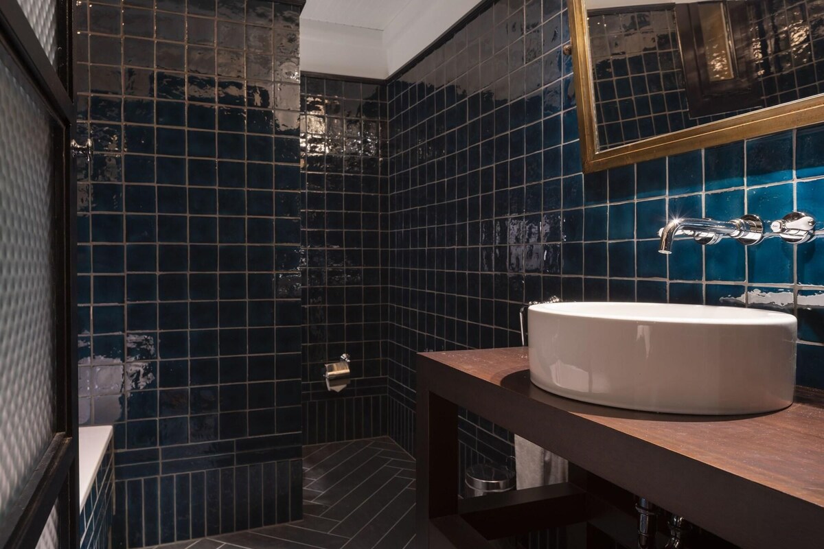 Uma casa de banho com azulejos azul escuro, uma pia branca e um espelho.