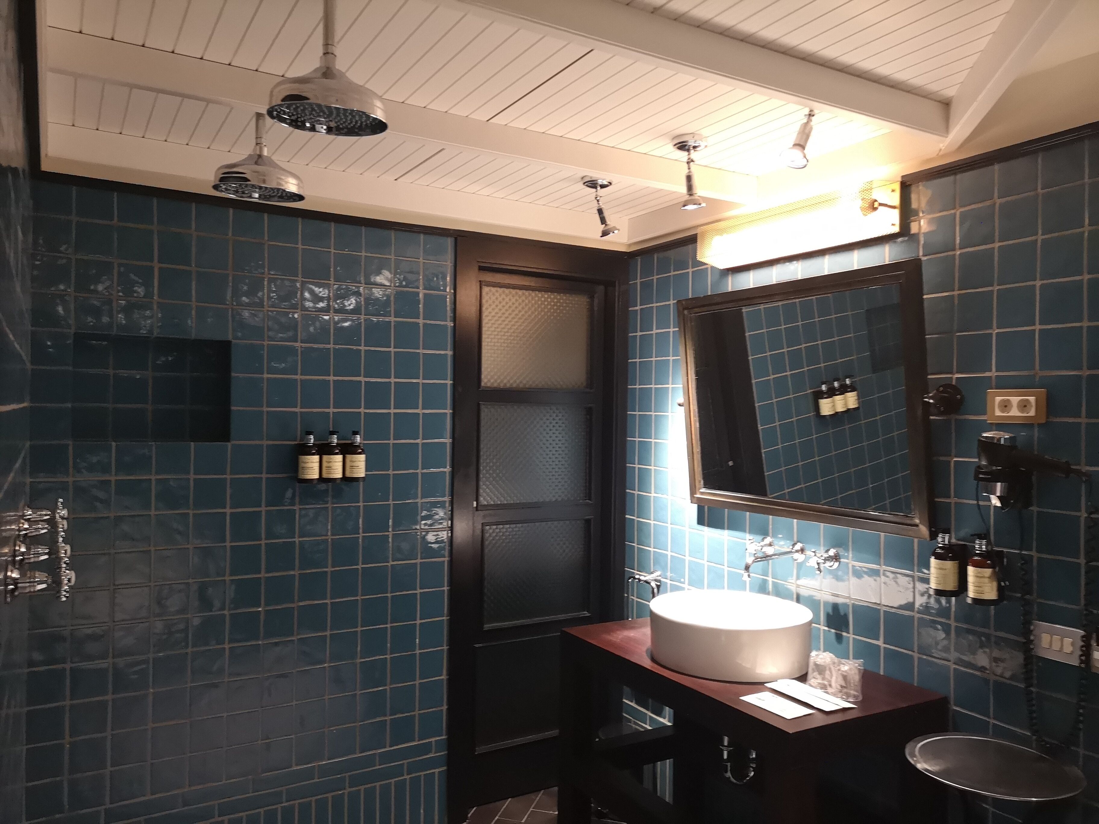 A bathroom with blue tiled walls, a wooden vanity, a white sink, and a mirror. There are ceiling lights and a door.